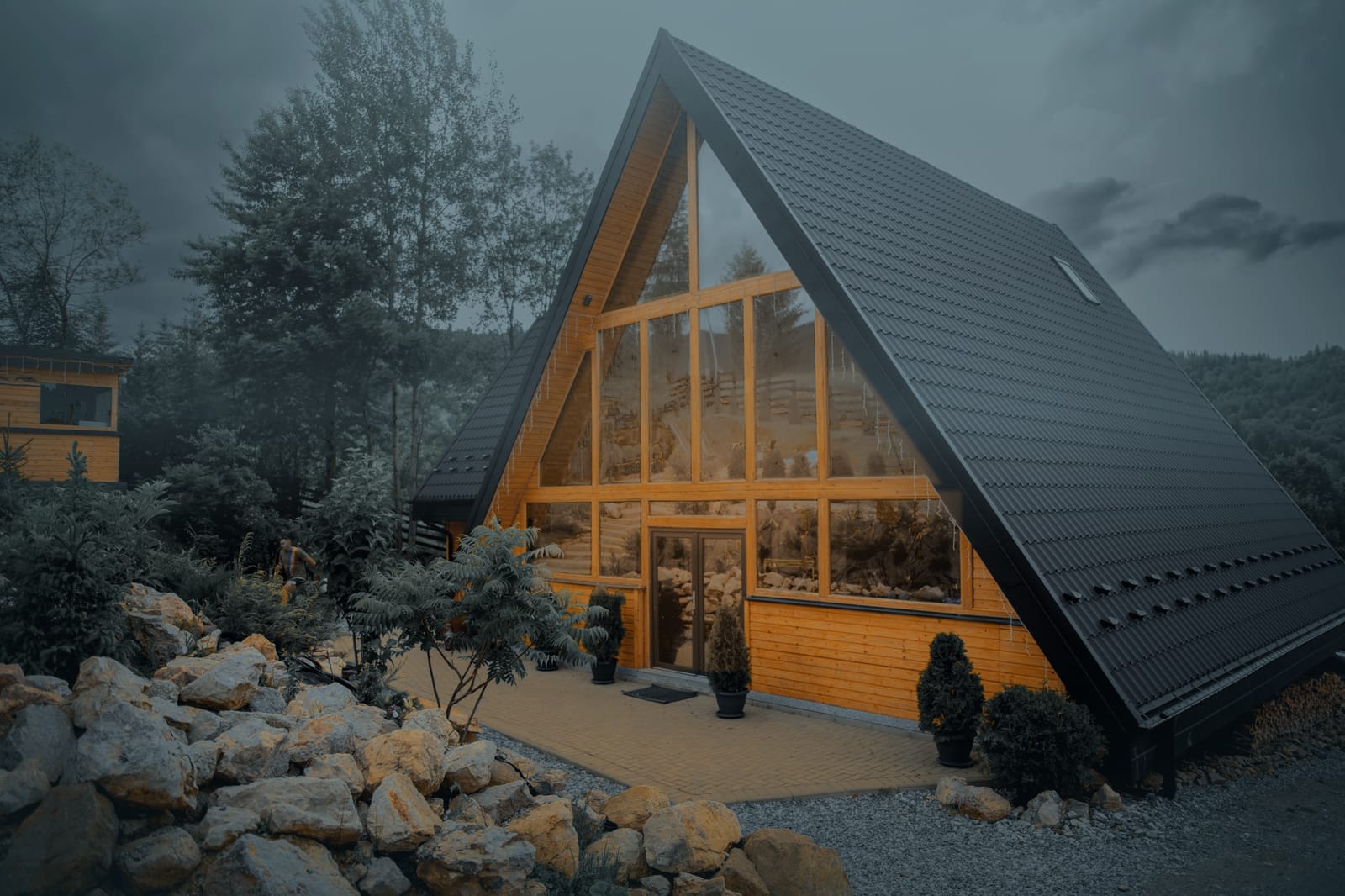 Cabana A-Frame Green View Rarău la amurg, cu lumină caldă în fațada de sticlă, între brazi și grădina cu pietre